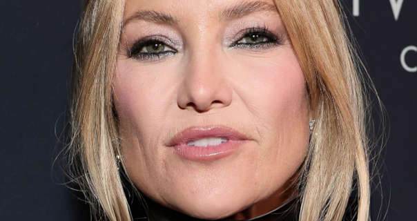 Kate Hudson wears her blonde hair tied back in a ponytail and with strands around her face. She stands in front of a dark grey event wall that has white text on it. Kate stars in a matt foundation, pink blouse, mascara, black eyeliner, light gloss lipstick and a pair of diamond earrings.