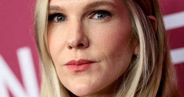 Lily Rabe is seen with her long blonde hair straightened and loose framing her face. She stands in front of a dark pink event wall that has white text across it. Lily wears a soft foundation with black mascara, gemstone earrings and a red lipstick.