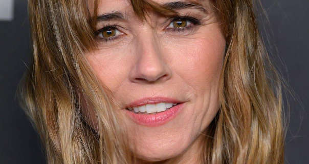 Linda Cardellini is seen with her hair loose, slightly curled and blown in the wind. She stands in front of a dark grey event wall with lighter grey text on it. Linda is seen in a soft foundation, blush, mascara and a light pink lipstick.