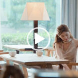 Zhao Lusi leans down and to the side as she puts away a gift. She sits at a light wood cafe table in front of Wan Peng with large windows all around. Zhao has her hair loose but pinned back. She wears a gold top with pearl earrings and a pink lipstick.