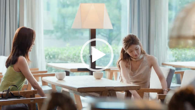 Zhao Lusi leans down and to the side as she puts away a gift. She sits at a light wood cafe table in front of Wan Peng with large windows all around. Zhao has her hair loose but pinned back. She wears a gold top with pearl earrings and a pink lipstick.