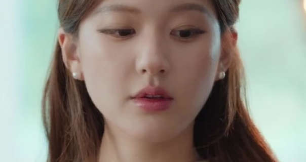 Zhao Lusi is seen with her light brown down and pinned back. She wears foundation with pink lipstick and a pair of pearl earrings. Zhao looks downwards and to one side. She appears in a large room in front of windows with daylight streaming in.