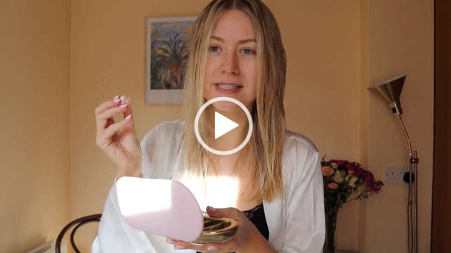 Meg Says stands in front of a small dark wood table and a pastel orange wall. She wears her long blonde hair loose and a little damp from washing. Meg holds her light pink and gold medicine case on one hand and a single pill in another. She is seen in a white dressing gown and a black sleepwear top.