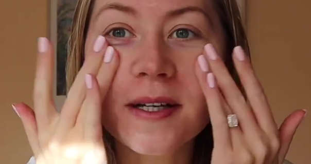 Meg Says is seen in front of a dark orange wall in her home. She has her long blonde hair loose and slightly damp from the shower. Meg touches her face with both of her hands. She is seen in no makeup and light pink nails.