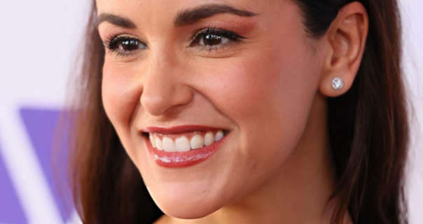 Melissa Fumero smiles widely and turns her head to the side. She appears in front of white and light purple wall at an event. Melissa has her long hair loose and pushed back. She appears in a matt foundation, blush, mascara, red gloss lipstick and a pair of large gemstone stud earrings.