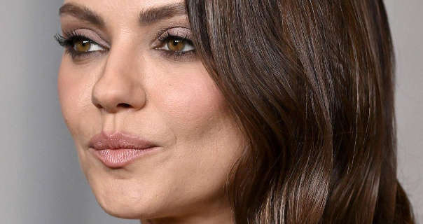 Mila Kunis is seen against a light grey backdrop at an event. She wears her long dark brown hair loose and in waves. She is seen from one side as she looks forwards. Mila wears a natural foundation with light blush, mascara and a pink lipstick.