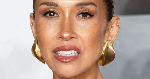 Myleene Klass stands in front of a white and grey movie event wall. She wears her hair slicked back in a tight bun. Myleene appears in a matt foundation, mascara, dark pink lipstick and a pair of large gold earrings.