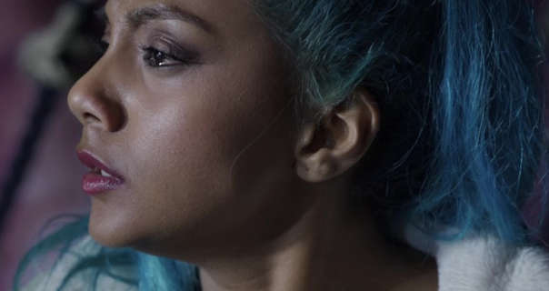 Charithra Chandran is seen looking into a bright light. She has her long hair tied up in a high ponytail and coloured blue. Charithra wears a soft blush with black mascara and a dark red lipstick.