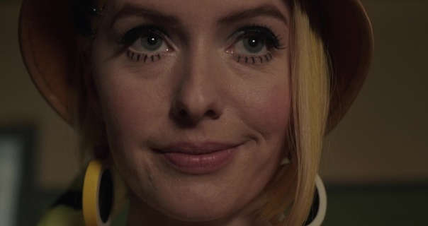 Jazzara Jaslyn smiles a little and glances forwards. She wears her blond hair pushed to one side with straightened strands framing her face. Jazzara wears a wide brim hat with plastic yellow hoop earrings, blush, exaggerated mascara and a dark pink lipstick.