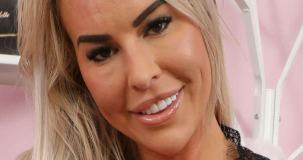 Luci Jones looks into the camera as she sits in front of a light pink wall and white metal shelves. She has her long thin blonde hair down and styled naturally. Luci is seen in a heavy foundation, blush, mascara and a pink gloss lipstick.