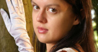 Kelsey is seen outdoors in a forest in dappled sunlight. She wears her long hair straightened and loose. Kelsey is seen in a pair of cream opera gloves, mascara, matt foundation and a pink lipstick. She touches the light trunk of a large tree with one hand as she gazes into the camera.