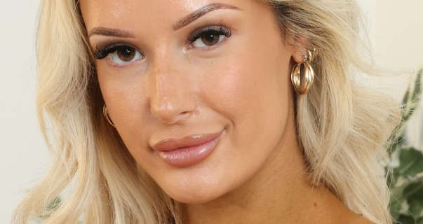 Paige F is seen with her light blonde hair down and in loose waves. She wears a thick matt foundation with mascara, dark pink gloss lipstick and a pair of thick gold hoop earrings. Paige appears in front of a cream wall with a small green leaf potted plant to one side.