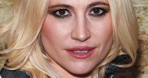 Pixie Lott stands in front of a worn sand colour brick wall. She has her blonde hair long and in soft waves. Pixie wears a black outfit with, blush, foundation, mascara and a dark pink lipstick.