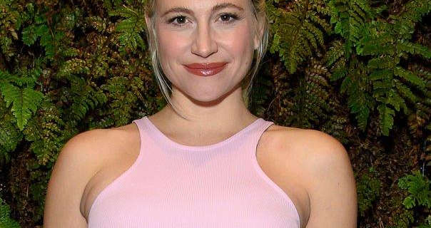 Pixie Lott stands in front of a green fern wall at an event. She has her long blonde hair tied up and back. Pixie wears a pink sleeveless dress with blush, mascara and a dark red lipstick.