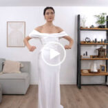 Roxxsaurus wears a long white off the shoulder ASOS gown. She appears in her YouTube filming set with her long brown hair tied back in a bun. Roxi holds her arms upwards to demonstrate the tight fit of the dress.