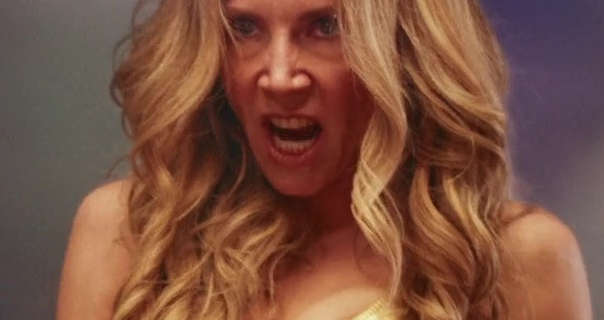 Sarah Chalke is seen with her long blonde hair in layered waves. She snarls and looks forward aggressively. Sarah stars in light makeup and a gold top.