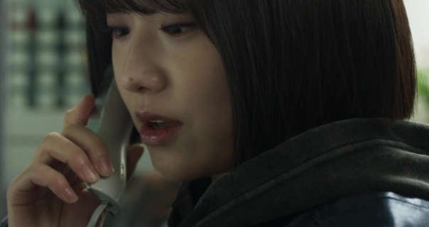 Kang Mi-na holds a small cream landline phone to her ear as she speaks. She wears a padded jacket with mascara, foundation and pink lipstick. Kang Mi-na sits in a slightly dark open plan office area.