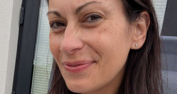 Vana takes a selfie photo for OnlyFans. She sits outside on a long grey chair in front of a patio style door. Vana is seen with her light brown hair loose and pushed back. She wears a pair of stud gemstone earrings with no makeup.