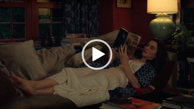 Rachel Weisz lies on a couch reading a paperback book in lamplight. She is seen in a red wall living room near a small plant, coffee table and in front of a row of wood frame windows. Rachel wears a long cream nightdress with a light and dark blue patterned dressing gown. She has her brown hair loose and in natural layers.