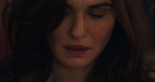 Rachel Weisz looks downwards with her eyes slightly closed and an intense look. She has her brown hair loose and in soft waves. Rachel is seen in a dark room. She appears in blush, mascara and a red lipstick.