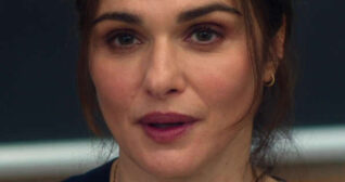 Rachel Weisz sits in front of a long silver frame blackboard. She wears her light brown hair tied back with some strands pulled down around her face. Rachel is seen in a light foundation, blush, mascara, dark pink lipstick and a pair of small gold earrings.