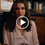 Rachel Weisz sits with her back against a dark wooden cabinet with books and a cardboard box on top of it. Rachel has her brown hair loose and fairly straight. She wears a cream top with gold jewellery, dark lipstick and matt foundation.