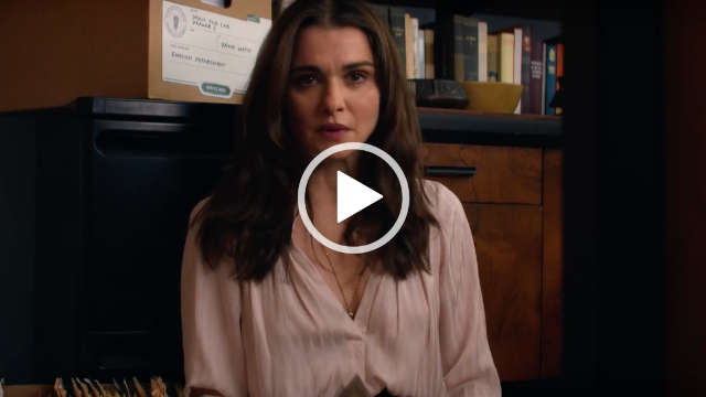 Rachel Weisz sits with her back against a dark wooden cabinet with books and a cardboard box on top of it. Rachel has her brown hair loose and fairly straight. She wears a cream top with gold jewellery, dark lipstick and matt foundation.