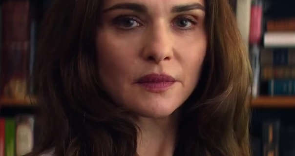 Rachel Weisz is seen with her long light brown hair loose and curled in gentle waves. She stands in front of wood bookshelf. Rachel stars in a matt foundation, light blush, mascara and a dark red lipstick.