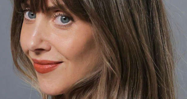 Alison Brie is seen in against a light grey backdrop. She wears her brown hair loose and with soft blonde highlights and a fringe. Alison stars in a matt foundation, curled lashes and a red lipstick.