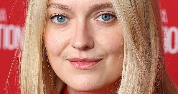 Dakota Fanning smiles slightly as she looks directly into the camera with her blue eyes. She is seen in front of a red wall with white event writing on it. Dakota has her long blonde hair down and straightened. She wears a matt foundation with a little mascara and a dark pink lipstick.
