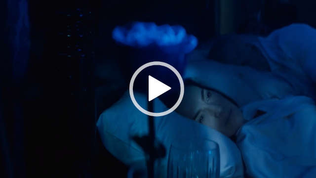 Ayelet Zurer lies in bed in the dark with her head on a large white pillow. She looks to the side in thought. Ayelet wears a long white dressing gown with her hair loose. A blue single stem rose and a small drinking glass are in the foreground.