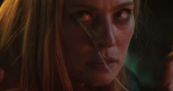 Deborah Ann Woll looks up and to the side with a serious and uneasy expression. She is outdoors at night with a spotlight hitting the top of her head. Deborah has her long ginger hair straightened and loose. Deborah wears a natural foundation with mascara and a dark pink lipstick.