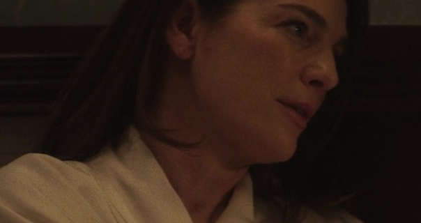 Ayelet Zurer is seen from the side leaning against a dark brown headboard at night. She wears her long brown hair loose and straightened. Ayelet stars in light makeup and a long dressing gown.
