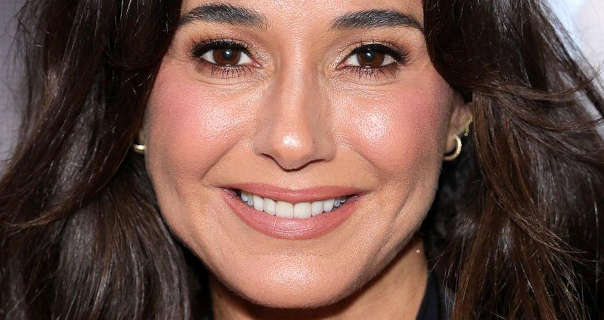 Emmanuelle Chriqui smiles as she appears in front of an event wall. She has her long, thick black hair loose and volumised in layers. Emmanuelle wears a dewy foundation with blush, mascara, dark pink eyeshadow, matt pale pink lipstick and a pair of small gold hoop earrings.