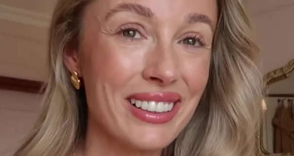Josie Irons smiles while standing near a closed door in a pink and white living room at home. She is seen next to a large gold frame antique mirror. Josie wears her blonde hair loose and in gentle waves. She appears in a matt foundation, curled lashes, blush, pink gloss lipstick and a pair if gold earrings.