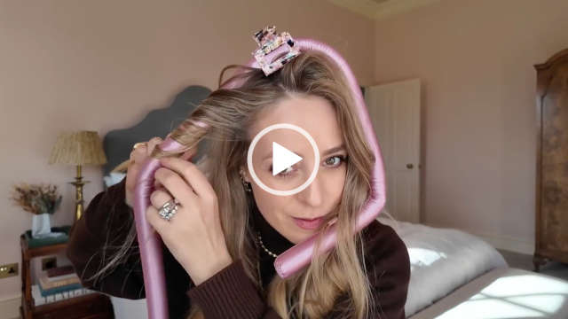 Fleur De Force is seen wrapping her hair around a pink hair rod to get heatless curls. She sits in a dusky pink and white bedroom at her home. Fleur appears in a matt foundation, mascara, dark pink lipstick. She wears a brown thin knitted jumper and gold jewellery.