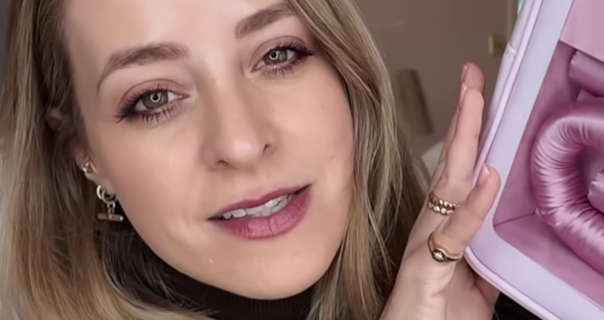 Fleur De Force is seen with her dark blonde hair loose and slightly curled. She sits in one of her neutrally decorated bedrooms at her home. She is seen in gold jewellery, matt foundation, mascara and a deep red lipstick. Fleur holds up a new heatless curling set in a bright pink colour.