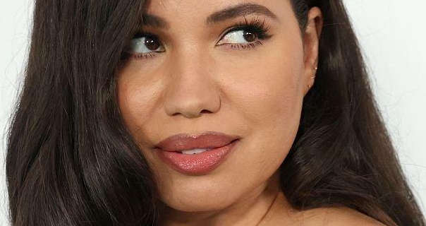 Jurnee Smollett wears a matt foundation with black mascara, pink eye shadow and a dark pink lipstick. She wears her thick black hair loose and in soft waves. Jurnee stands in front of a dark white wall and looks upwards and to the side.