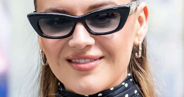 Kelsea Ballerini is seen outdoors on a city street in the day. She wears her long light brown hair loose and tied back. Kelsea appears in a pair of thick black frame sunglasses, foundation, dark pink lipstick, gold earrings and a black and white high neck spotty dress.