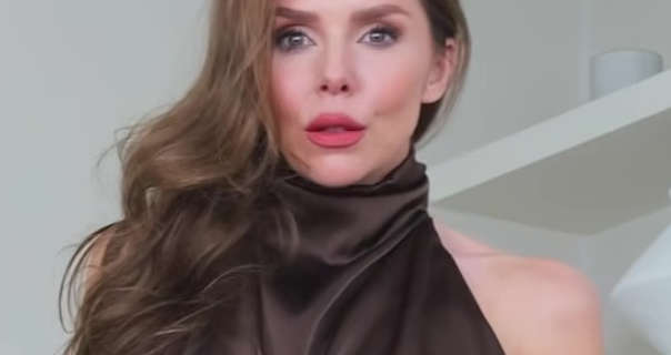 Laura Blair appears in front of a white wall and some long white wood display shelves. Laura has her brown hair down, pushed to one side and in layered waves. She wears a brown halter neck dress with foundation, blush, mascara and a red lipstick.