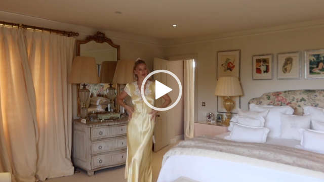 Leonora Smee is seen at a bit of a distance as she stands in her large filming bedroom at her countryside house. She looks to one side as she runs both her hand up the light yellow lace trim dress she is wearing. Leonora has her long blonde hair tied back with a scrunchie.