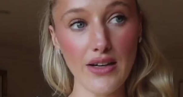 Leonora Smee looks to the side as she stands in her filming bedroom at home. She wears her long long hair loose and pushed back. Leonora appears in a red blush, matt foundation, mascara, pink lipstick and a pair of small gem earrings.