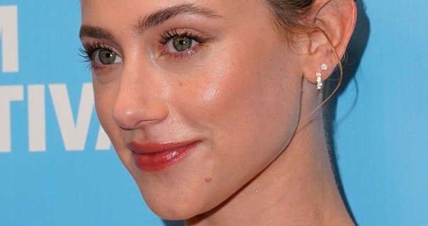 Lili Reinhart is seen with her blonde hair tied up in a slick bun. She stands in front of a light blue wall that has dark white text on it. Lili wears a soft foundation with mascara, red lipstick and a few small diamond studded earrings.