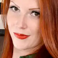 Ruby looks into the camera and smiles. She has her long red hair loose and straightened. She appears in front of a brown wall with a large abstract painting hung on it. Ruby wears a matt foundation with a bright red lipstick and black mascara.