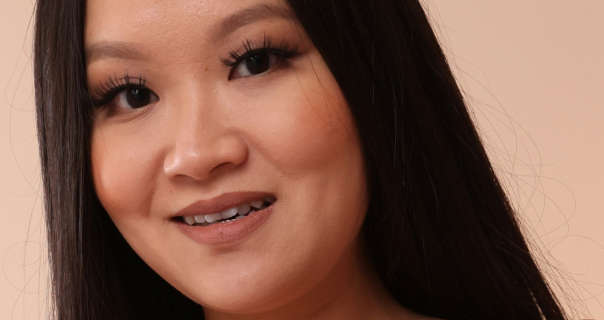 Ophelia Spice is seen in front of a pastel orange wall. She wears her long black hair straightened and loose. Ophelia appears in a soft foundation, black mascara, light blush and a dark pink matt lipstick.