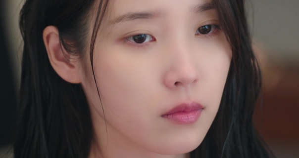 IU is seen with her long black hair loose and pushed a little to one side. She wears a light foundation with pink eye shadow and a pink lipstick. IU is seen in a dressing room during the day with as she gazes forwards in thought.