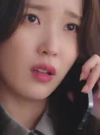 IU holds a silver frame mobile phone to her ear as she talks. She has her black hair loose and in soft waves. IU wears a grey blouse with a light foundation, mascara and pink lipstick.