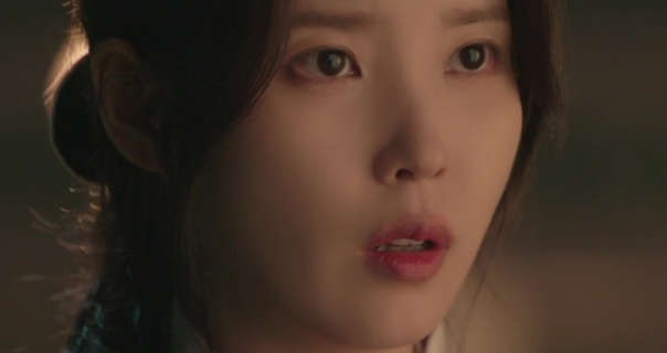 Lee Ji-eun is seen outdoors at night. She has her long black hair tied back in a messy bun. IU appears in a soft foundation and a light pink lipstick. She looks forwards with a concerned expression.
