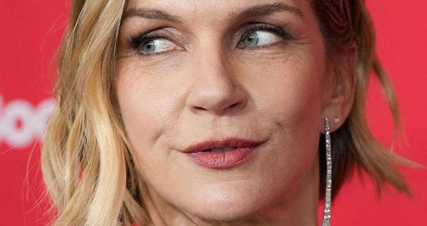 Rhea Seehorn is seen standing in front of a pastel red wall with dark white writing on it. She wears her thick blonde hair cut short and styled in waves. Rhea appears in a light coverage foundation, mascara, dark red lipstick and a pair of thin hanging diamond encrusted earrings. She looks to one side with a slight smile.