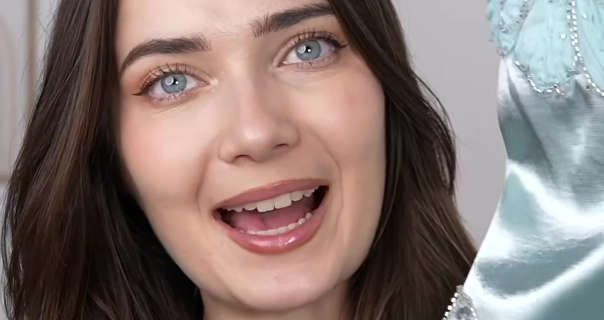 Roxxsaurus appears in front of a light taupe wall. She has her long brown hair loose and in natural waves. Roksana wears a matt foundation with pink eyeshadow, curled lashes and a dark pink gloss lipstick. She holds up a light blue gown from Shein.